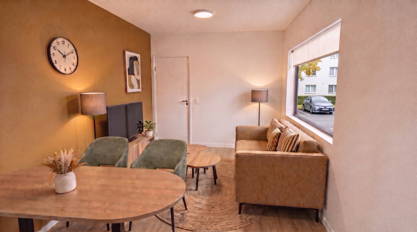 De woonkamer met een bank bij het raam, een televisie, een eettafel met stoelen en warme kleuren.